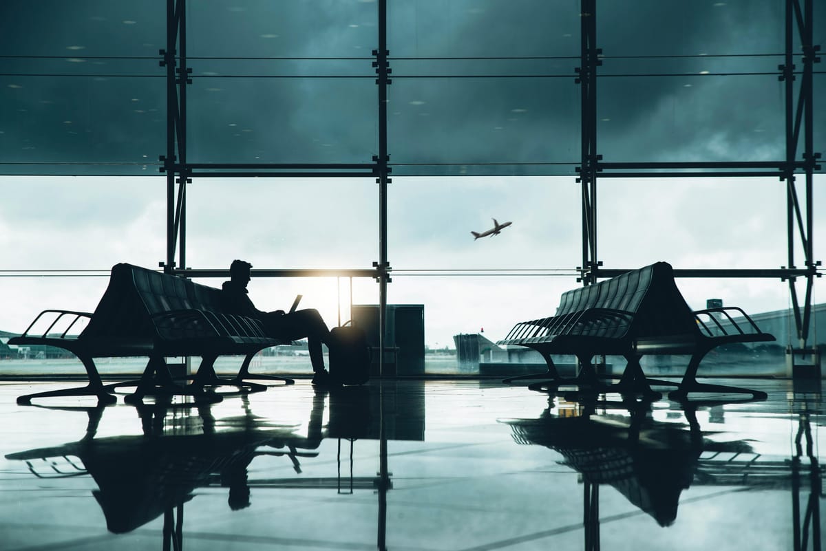 Reisender mit Laptop auf einer Bank im Flughafen-Terminal, neben sich ein Koffer, draußen startet ein Flugzeug