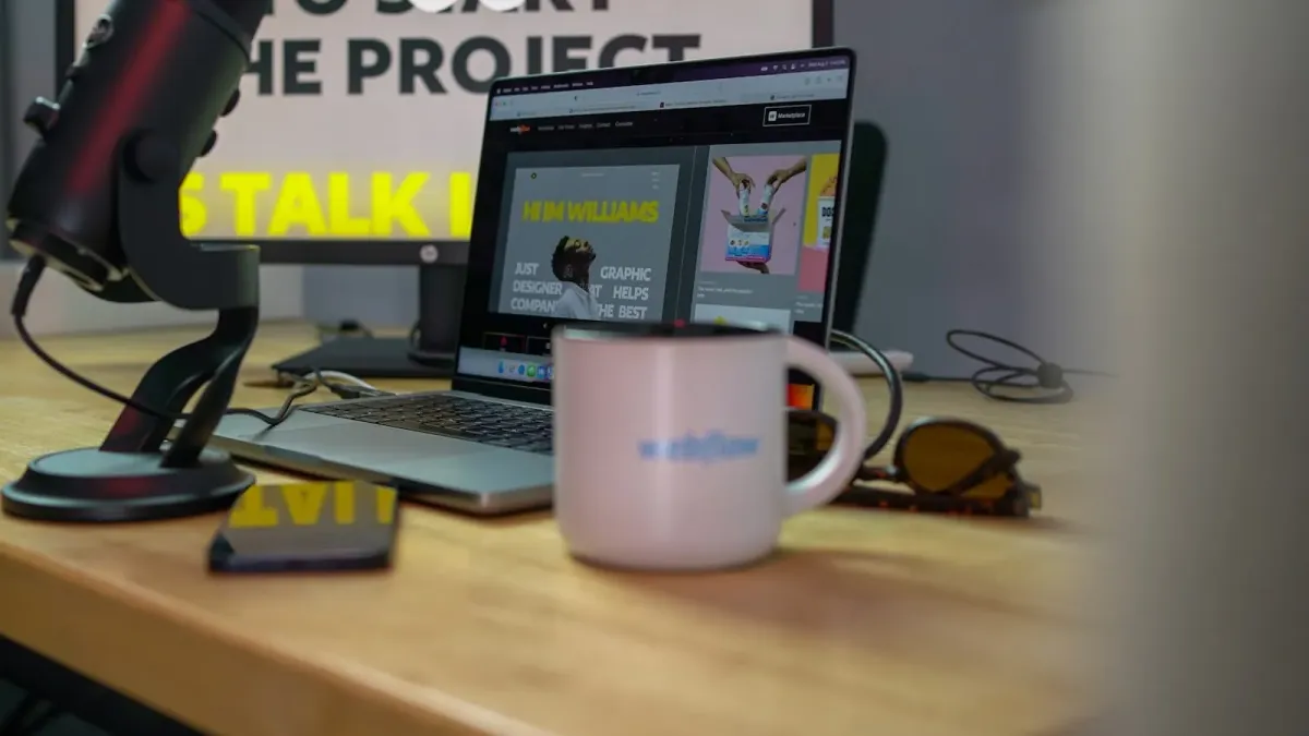 Laptop auf einem Holzschreibtisch, im Browser eine Portfolio-Website, daneben Mikrofon, Kaffeetasse und Sonnenbrille