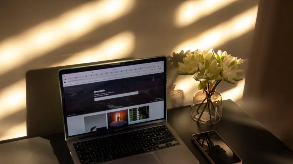 Aufgeklappter Laptop mit geöffneter Website im Browser auf einem Tisch, daneben Blumenvase und Smartphone im Abendlicht