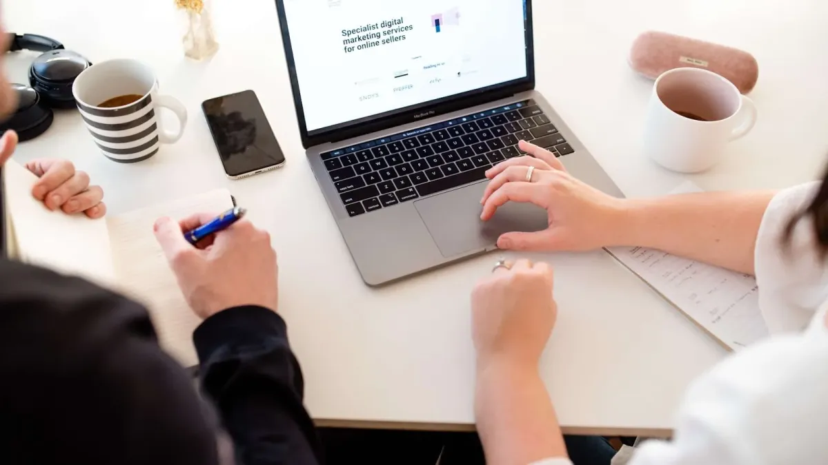 Zwei Menschen prüfen am Besprechungstisch eine Website auf dem Laptop, daneben Notizblock, Stift und Kaffee