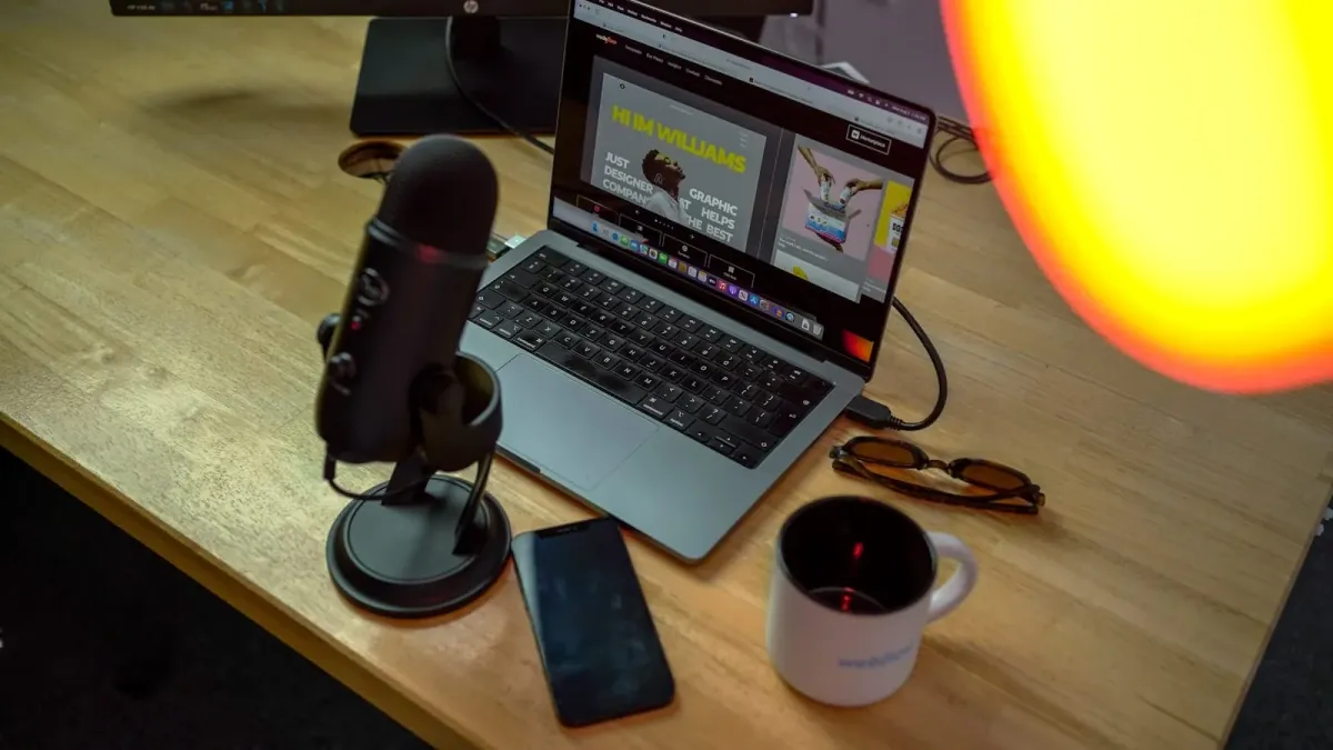 Aufgeklappter Laptop mit einer Website im Browser auf einem Holzschreibtisch, daneben Mikrofon, Smartphone und Tasse