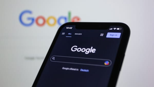 Smartphone mit der Google-Startseite im Dunkelmodus, dahinter unscharf das bunte Google-Logo