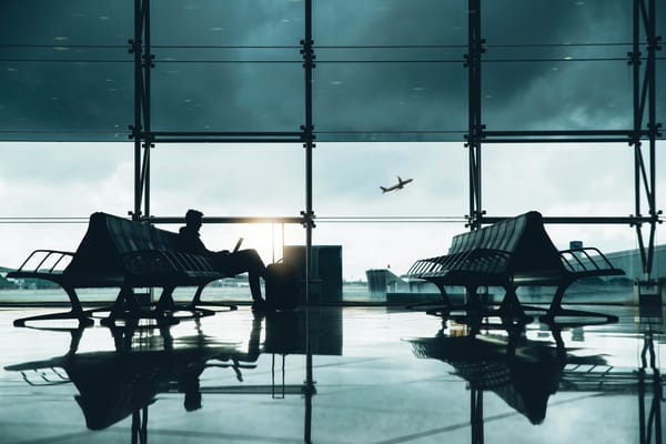 Reisender mit Laptop auf einer Bank im Flughafen-Terminal, neben sich ein Koffer, draußen startet ein Flugzeug