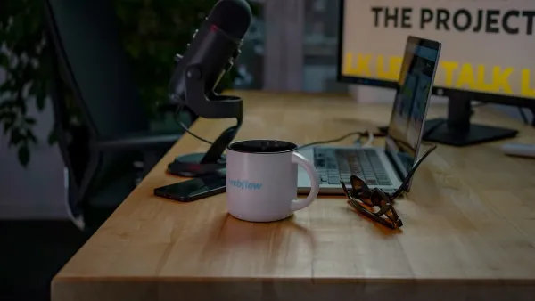 Weiße Tasse mit Webflow-Logo auf einem Holzschreibtisch, dahinter Podcast-Mikrofon, aufgeklappter Laptop und Brille