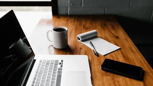 Aufgeklappter Laptop, Kaffeetasse, handschriftlich beschriebener Notizblock und Smartphone auf einem Holztisch