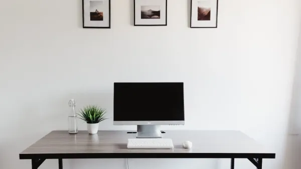 Aufgeräumter Schreibtisch mit ausgeschaltetem Monitor, weißer Tastatur und Maus vor einer weißen Wand mit drei Bilderrahmen