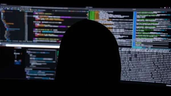 Dunkle Silhouette einer Person vor drei Monitoren voller farbigem Programmcode und Terminal-Ausgaben