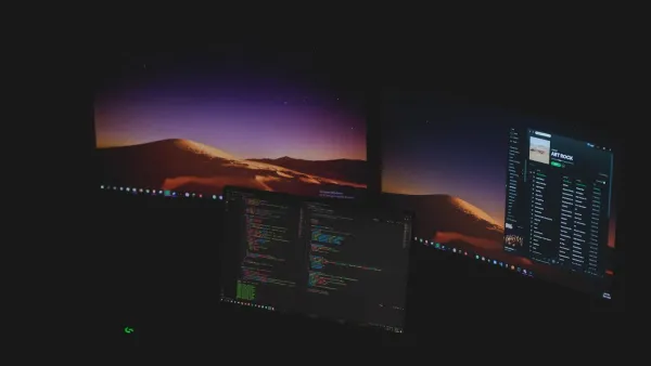 Drei Monitore in einem dunklen Raum, in der Mitte ein Code-Editor, auf den äußeren ein Wüstenmotiv als Hintergrund